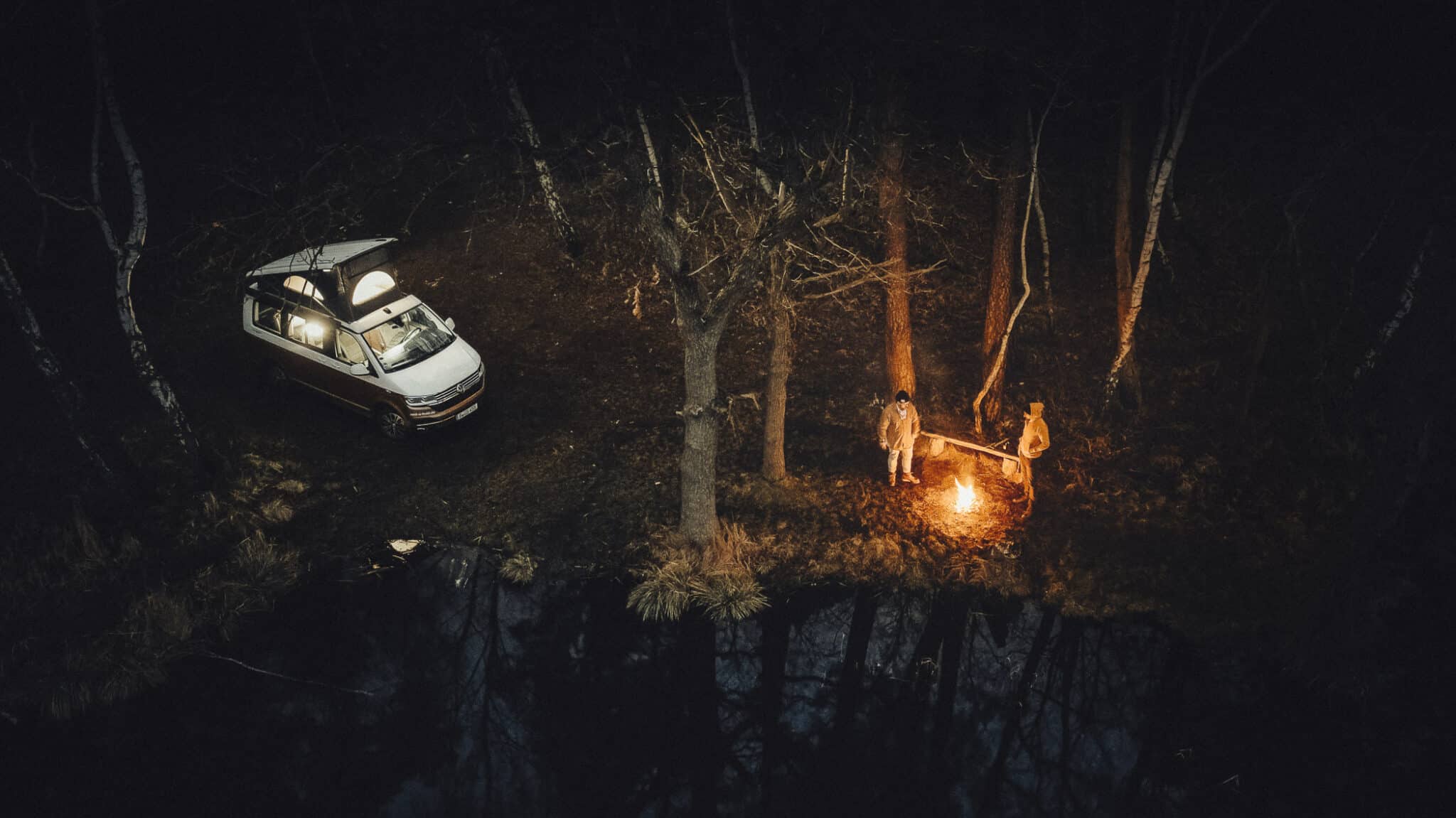 Unternehmensfotografie für einen Bulli-Vermieter aus Berlin: Key Visual mit VW Campingbus und Lagerfeuer am Seeufer bei Nacht, aus der Luft fotografiert